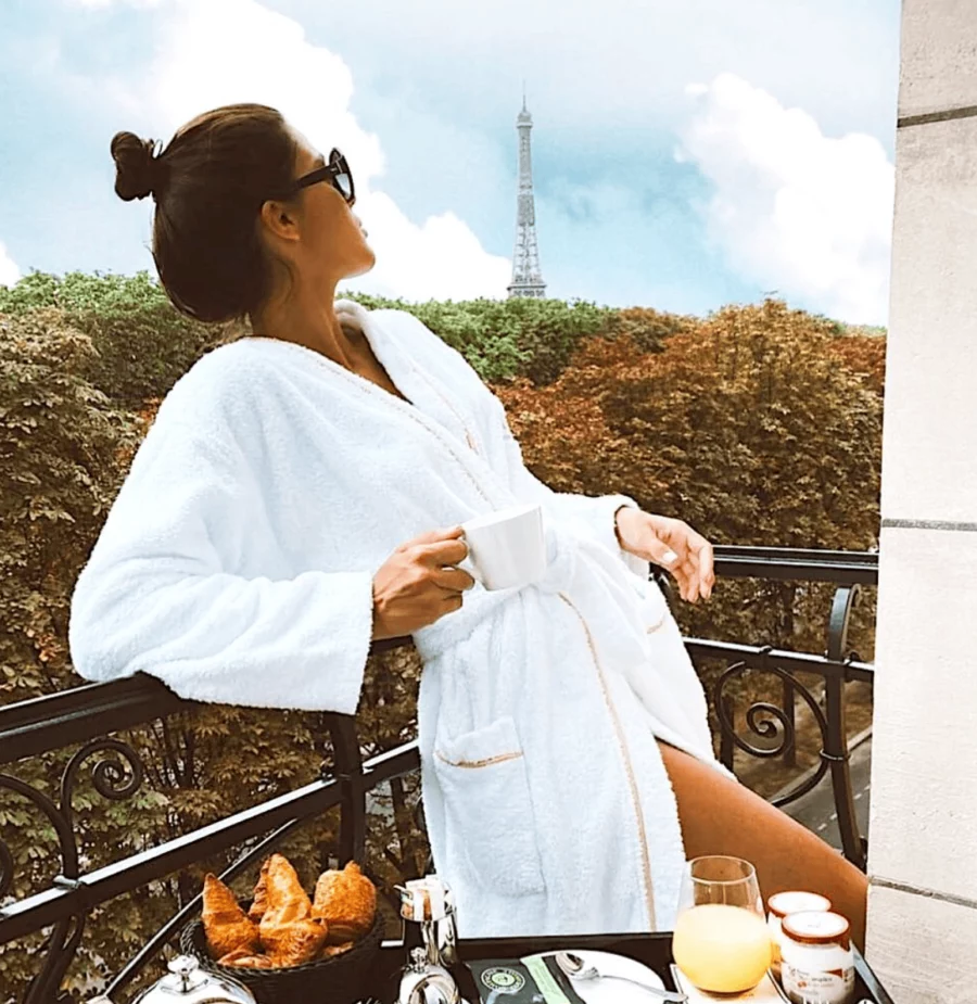 Femme sur un balcon d'hôtel 5 étoiles, vêtue d'un peignoir élégant, admirant une vue panoramique et luxueuse.