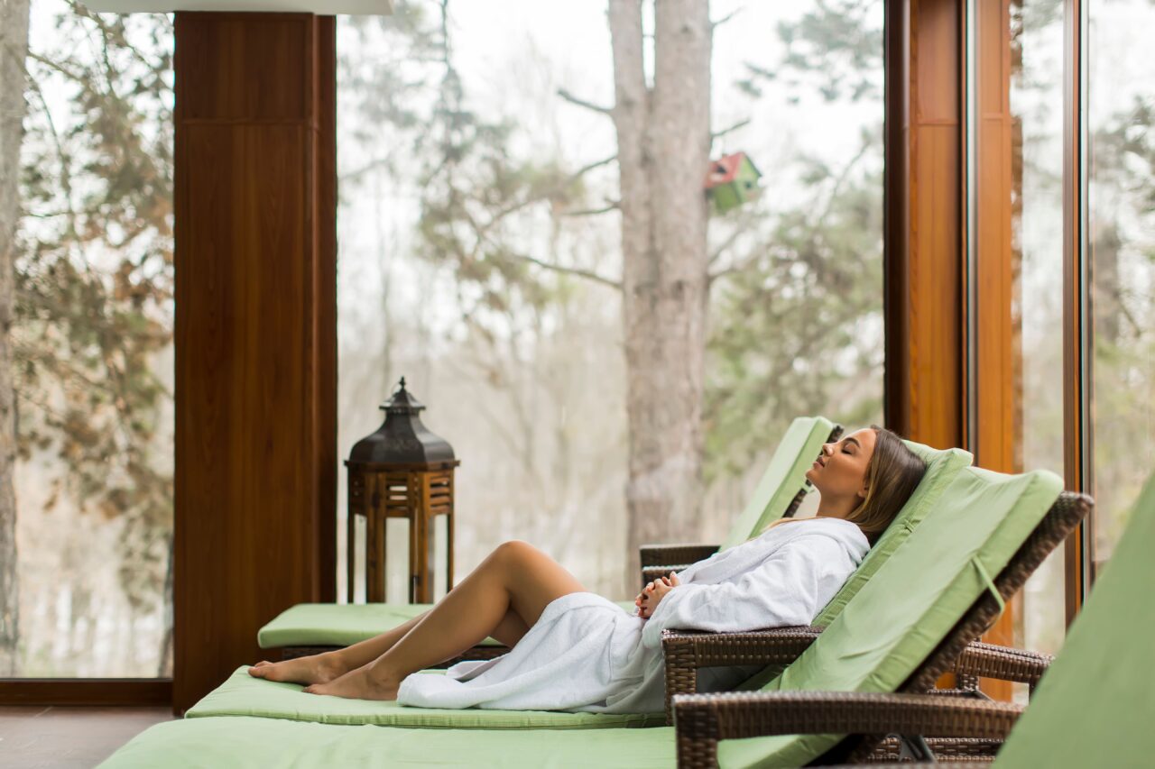 Une femme est allongée sur un fauteuil en bambou au bord de la piscine d'un spa, portant un peignoir. L'atmosphère est paisible, avec une lumière douce qui reflète sur l'eau, créant une ambiance de détente et de bien-être. Le mobilier en bambou ajoute une touche naturelle et exotique à l'ensemble, contribuant à un cadre de relaxation parfait.